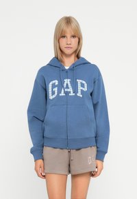 Blå hettegenser med glidelås og stor, lyseblå "GAP"-logo på forsiden. Den har en hette med snøring og frontlommer. Laget av myk bomullsmateriale.