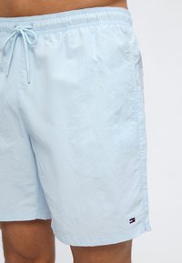 Ανοιχτό γαλάζιο μαγιό σορτς με ελαστική μέση και κορδόνι, φορεμένο από άτομο με εμφανή μαυρισμένα πόδια.