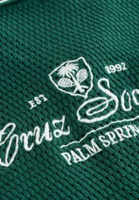 Tissu vert texturé avec un motif en mesh, présentant des graphismes brodés blancs : palmier et texte 'Palm Springs'. L'année '1992' est incluse.