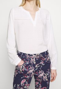 Blouse blanche à manches longues avec un col en encoche et un motif texturé, associée à un pantalon bleu foncé avec un imprimé cachemire coloré.