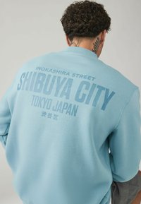 Svetlomodrý sveter s výrazným textom "SHIBUYA CITY TOKYO JAPAN" a "INOKASHIRA STREET", vyrobený z mäkkej, textúrovanej látky.