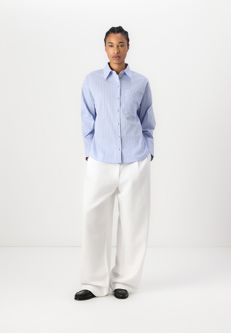 Niebanalna, niebiesko-biała koszula z paskami zapinana na guziki z kieszonką, zestawiona z luźnymi białymi spodniami i czarnymi butami typu slip-on. Prosty, dopasowany krój.