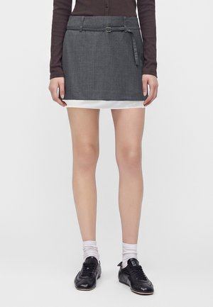 Femme portant une mini-jupe gris foncé ceinturée, un haut à manches longues foncé, des chaussettes blanches au niveau des chevilles et des chaussures noires à lacets, debout sur un fond uni.