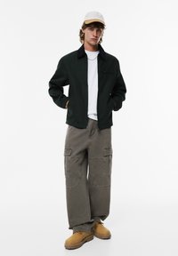 Donkergroene zip-up jas met zwarte kraag, witte t-shirt, grijze cargobroek met meerdere zakken en stevige tan laarzen. Witte pet gedragen.