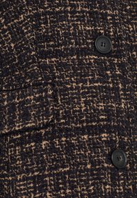 Tela de chaqueta texturizada en negro y beige con un patrón tejido; incluye un solo botón negro y un detalle de bolsillo con solapa.