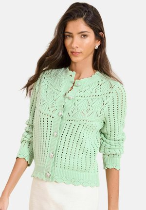 Kvinde iført en lysegrøn strikket cardigan med hjerteformede knapper og bølgede kanter, kombineret med et hvidt skørt.