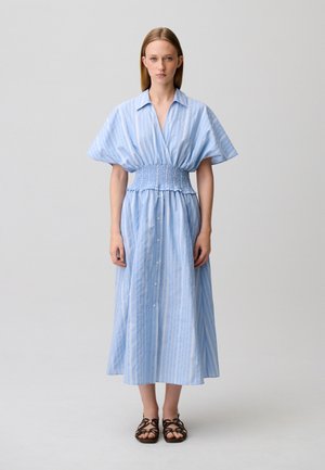 Donna in piedi di fronte, indossa un vestito midi a righe azzurro chiaro con bottoni, maniche ampie e vita arricciata, abbinato a sandali marroni.