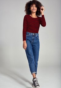 Maglietta a maniche lunghe a righe rosse e nere abbinata a jeans in denim blu a vita alta e sneakers nere alte, su uno sfondo grigio chiaro.