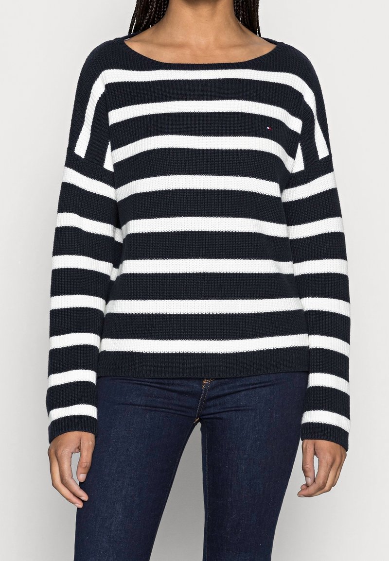Femme portant un pull en maille à rayures horizontales bleu marine et blanches avec un jean bleu foncé, debout devant un fond uni.
