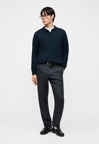 Άνδρας με μαύρα μαλλιά και γυαλιά φοράει μπλε πόλο με μακριά μανίκια, σκούρα γκρι παντελόνια, μαύρη ζώνη, λευκές κάλτσες και μαύρα επίσημα παπούτσια.