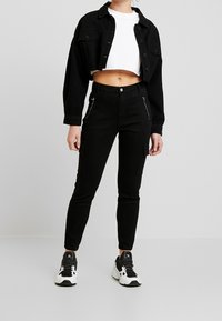 Chaqueta de mezclilla negra recortada con cierre de botones y bolsillos en el pecho, combinada con pantalones cargo negros de cintura alta con bolsillos con cremallera. Camiseta blanca debajo.