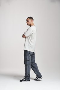 Tricou alb din bumbac, croială lejeră. Pantaloni cargo gri cu multiple buzunare. Adidași negri și albi. Poziție în picioare cu brațele încrucișate.