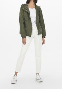 Olijfgroene lichtgewicht jas met een capuchon, elastische manchetten en een rits aan de voorkant, gecombineerd met witte rechte broek en witte sneakers.