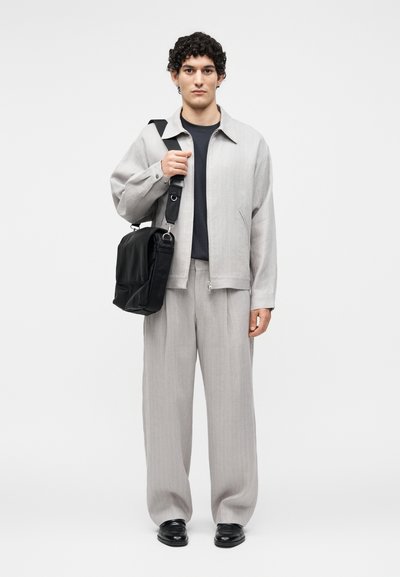 Νέος άνδρας στέκεται, φοράει ανοιχτό γκρι κουστούμι, μαύρο πουκάμισο, μαύρα παπούτσια και κρατάει μαύρη τσάντα ώμου με φαρδιά ζώνη.