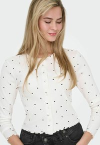 Cardigan blanc côtelé avec pois noirs, col rond, fermeture à boutons et détails à volants sur l'ourlet. Manches longues complètent le modèle.