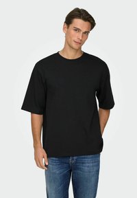 Schwarzes T-Shirt mit kurzen Ärmeln aus Baumwollstoff, locker geschnitten, mit rundem Halsausschnitt, getragen zu blauen Denim-Jeans. Einfaches Design in unifarbener Optik.