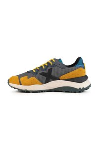 Zapatilla deportiva con parte superior de tela gris, acentos de gamuza amarilla, logotipo negro y detalle en teal en el talón. Presenta una suela de goma blanca y verde.