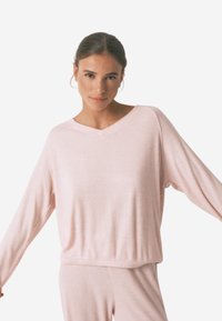 Lichtroze langemouwtop met een v-hals, ribbelstructuur en een ontspannen pasvorm. Zachte stof met verlaagde schouders en een cropped lengte.