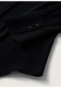 Poignet de chemise noire à manches longues avec deux boutons noirs sur un tissu lisse, posé à plat sur une surface blanche.