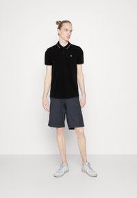 Sort polo t-shirt med hvid kant og logo, parret med mørke shorts. Modellen har lysegrå sneakers på. Simpelt design og afslappet pasform.