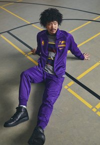 Fato tracksuit roxo dos Lakers com logo e ténis Nike pretos, sentado numa quadra de basquetebol com linhas amarelas e pretas.