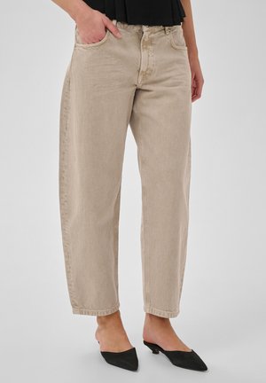 Persona che indossa jeans beige larghi, top nero senza maniche e scarpe nere basse senza lacci, mano in tasca, vista ritagliata.