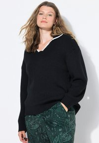 Vrouw met golvend haar, gekleed in een zwart geribbeld sweater en groene paisley-gepatroneerde broek, staande met één hand in de zak tegen een witte muur.