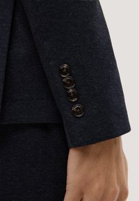 Ärmelende eines marineblauen Anzugsakts mit einer strukturierten Stoffstruktur, versehen mit vier dunklen Knöpfen entlang der Kante und einem glatten Finish am Ärmel.