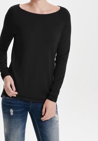 Maglietta nera a maniche lunghe con scollo ampio e una texture liscia, abbinata a jeans blu strappati con accenti scoloriti e buchi.