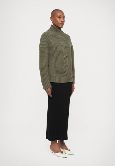 Suéter de malha canelada verde-oliva com gola alta, apresentando um padrão central de malha de cabos e um corte descontraído. Combinado com uma saia preta e sapatos brancos.