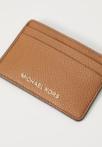 Rudas odinis kortelių laikiklis su tekstūruotu paviršiumi, turintis keturis kortelių skyrelius ir auksu įspaustą "MICHAEL KORS" logotipą priekyje.