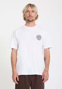 Homme blond portant un t-shirt blanc avec un petit dessin de soleil noir et du texte sur le côté gauche de la poitrine, debout devant un fond blanc uni.