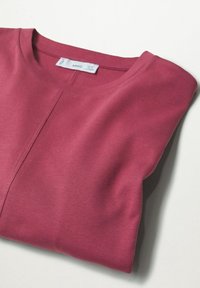 Chemise à manches longues rose vieux rose pliée avec encolure ronde, couture centrale visible et étiquette de marque blanche à l'intérieur du col sur fond blanc.