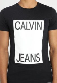 Zwart T-shirt gemaakt van katoen met een ronde hals, voorzien van een grote witte rechthoekige grafiek met de tekst "CALVIN JEANS" in vetgedrukt zwart. 