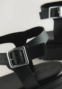 Svarta lädersandaler har en dubbelremdesign med en silverfärgad rektangulär spänne. Sulan är texturerad och något upphöjd för komfort.