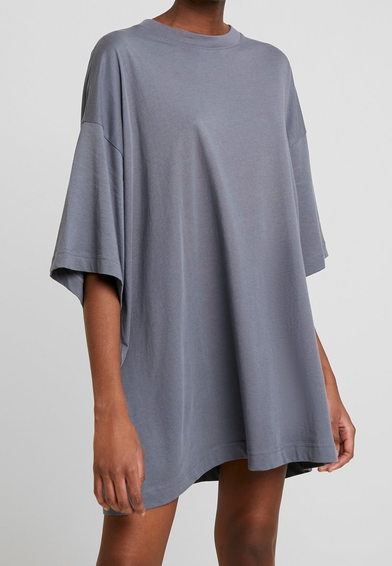 Person iført en løs, oversized grå kortærmet T-shirt kjole, der går til midt på låret, med afslappede sænkede skuldre og rund halsåbning.