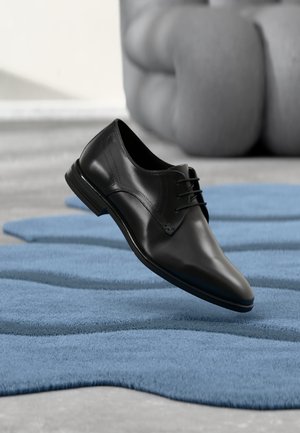 Schwarzer Lederschuh mit Schnürsenkeln, der auf einem blauen, strukturierten Teppich liegt, mit einem verschwommenen grauen Sofa im Hintergrund.