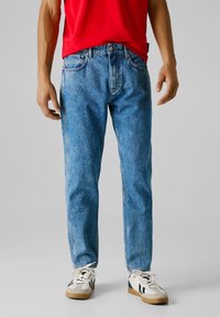 Persona vistiendo jeans de mezclilla azules, zapatillas blancas con franjas negras y una camisa roja, de pie contra un fondo gris liso.