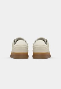 Une paire de baskets beiges avec une semelle marron vues de l'arrière sur un fond blanc.