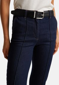 Tmavě modré riflové džiny s texturovaným povrchem, s kontrastním oranžovým švem, vysokým pasem a páskem s černým krokodýlím vzorem a stříbrnou sponou.