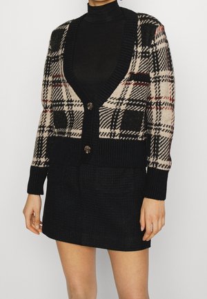 Kvinde iført sort rullekrave, sort og beige ternet cardigan med knapper og sort tekstureret nederdel, vist fra halsen til midt på låret.