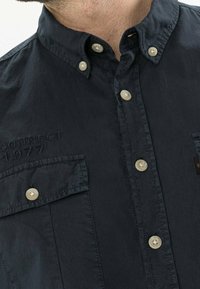 Dunkelblaues Button-Up-Hemd mit klassischem Kragen, Brusttaschen und gesticktem Detail "CONNECT 1977". Dezente Textur mit kontrastierenden Knöpfen.