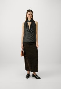 Top nero con collo a cowl e design a chiave, abbinato a una lunga gonna marrone scuro. Il modello tiene in mano una piccola borsa marrone e indossa mocassini neri.