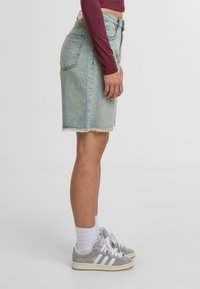 Persona vistiendo una falda de mezclilla azul claro con dobladillo deshilachado, un jersey de manga larga burdeos, calcetines blancos y zapatillas Adidas grises, de pie de lado.