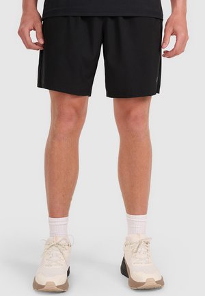 Piernas con pantalones cortos deportivos negros, calcetines blancos y zapatillas de correr beige con cordones sobre un fondo gris liso.