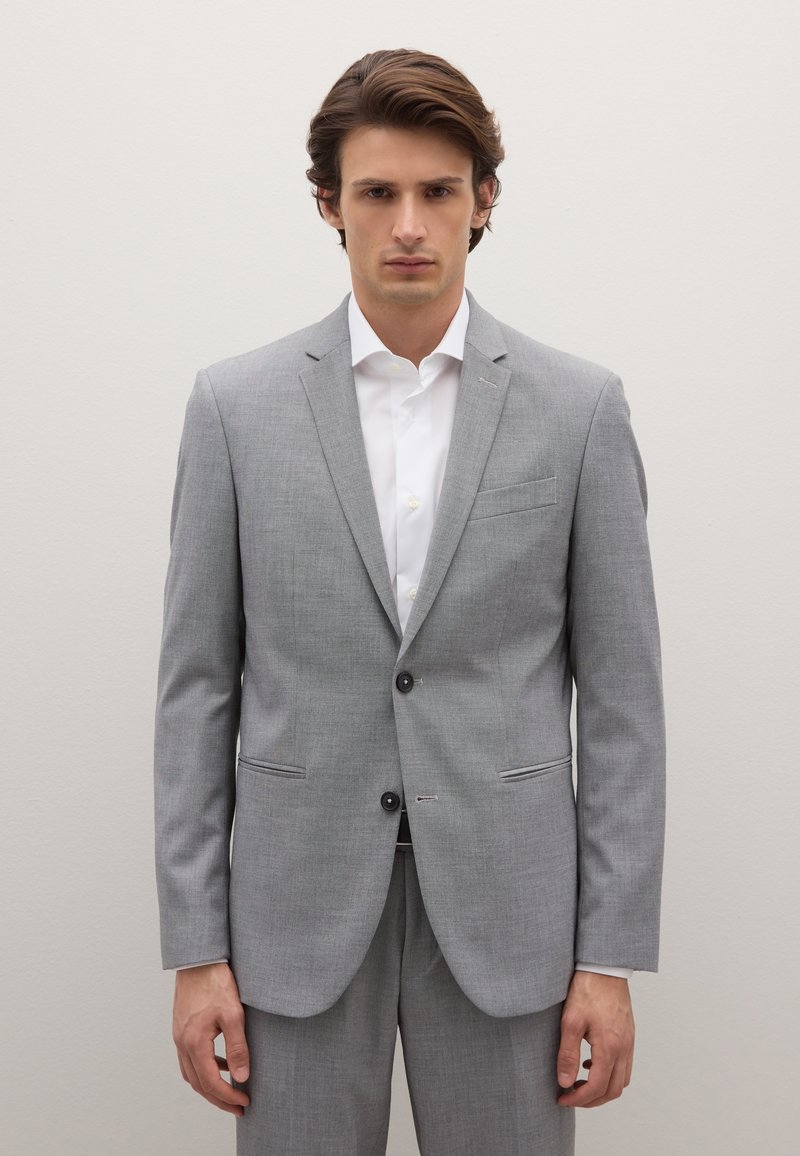 Lichtgrijs colbert van een gestructureerde stof, uitgevoerd met een revers met inkepingen, twee knopen en een borstzak. Draagt over een wit overhemd.