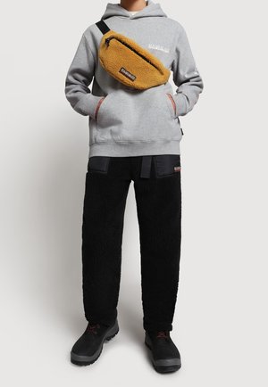 Person trägt grauen Kapuzenpullover, schwarze Fleece-Hose mit Taschen, schwarze Stiefel und eine beige Fleece-Umhängetasche quer über der Brust.