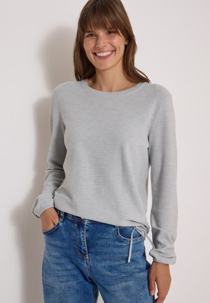 Femme souriante aux cheveux bruns portant un haut à manches longues gris clair texturé et un jean bleu, debout avec une main dans la poche sur un fond uni.