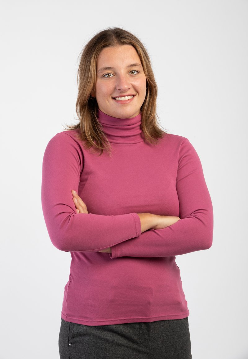 Maglietta a maniche lunghe rosa con collo alto, dal design aderente e dalla texture morbida, abbinata a pantaloni grigi scuri, con le braccia incrociate davanti.