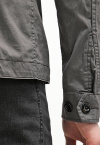 Szara bawełniana kurtka o lekko teksturowanej powierzchni, z mankietem na guziki i kontrastującymi, czarnymi guzikami, prezentowana z czarnymi spodniami jeansowymi.